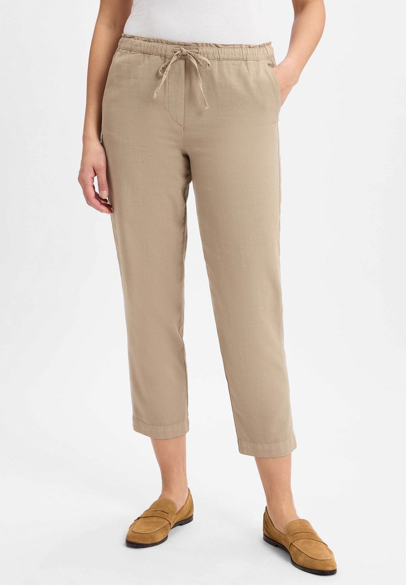 Femme portant un pantalon beige court à cordon de serrage et des mocassins en daim beige, se tenant avec une main dans la poche contre un fond blanc.