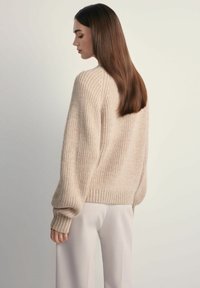 Beżowy, dziany sweter o luźnym kroju, z ribbowanym dołem i mankietami, oraz teksturowanym materiałem. Nosi się go na jasnych spodniach.