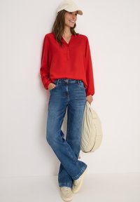Blusa rossa a maniche lunghe con scollo a V e bottoni abbinata a jeans blu svasati. Cappello beige e sneakers, mentre tiene una borsa color crema chiaro.