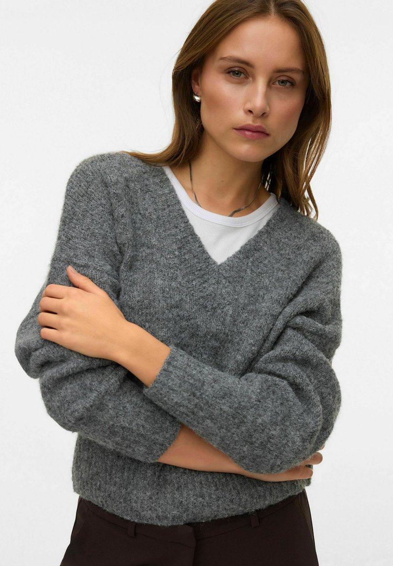 Grauer Strickpullover mit V-Ausschnitt und langen, leicht gepufften Ärmeln. Weiche Textur mit gemütlicher Passform, über einem weißen Oberteil getragen.