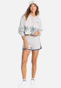 Vrouw in een grijze hoodie crop sweatshirt met palmbomenprints en bijpassende shorts, met witte sokken en een lichtgekleurde pet.