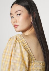 Blusa amarela de gingham com mangas bufantes, detalhe em franzido e design às costas aberto. Modelo usa brincos coloridos em forma de argola.