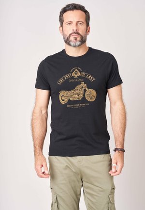 Man met baard die een zwart t-shirt draagt met een vintage motorfietsafbeelding en kaki cargobroek, staand tegen een effen achtergrond.