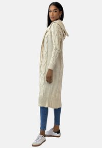 Stickad kofta i krämfärg med flätmönster, långa ärmar och huva. Kombinerad med blå jeans och vita sneakers med svarta detaljer.