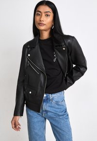 Chaqueta de cuero negra de motorista con herrajes plateados, cremallera asimétrica y bolsillo frontal. Combinada con una camiseta negra y vaqueros azules.