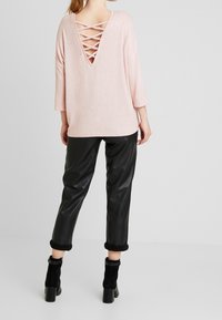 Jasnoróżowa, luźna bluzka z głębokim dekoltem na plecach i szczegółem w postaci krzyżujących się ramiączek, zestawiona z czarnymi spodniami z imitacji skóry i botkami do kostek.