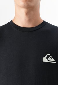 Un gros plan du cou et de la poitrine d'une personne portant un t-shirt noir avec un petit logo blanc sur le côté gauche.