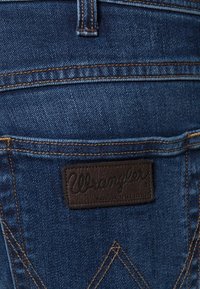Vaqueros de mezclilla azul oscuro con un distintivo parche de cuero Wrangler en el bolsillo trasero, con costuras dobles y una textura sutil.