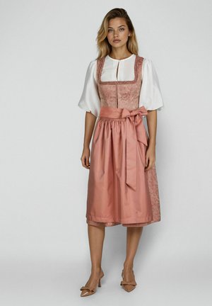 Jonge vrouw die een traditionele roze dirndl-jurk draagt met witte pofmouwen en een gebonden schort, staand tegen een effen achtergrond.