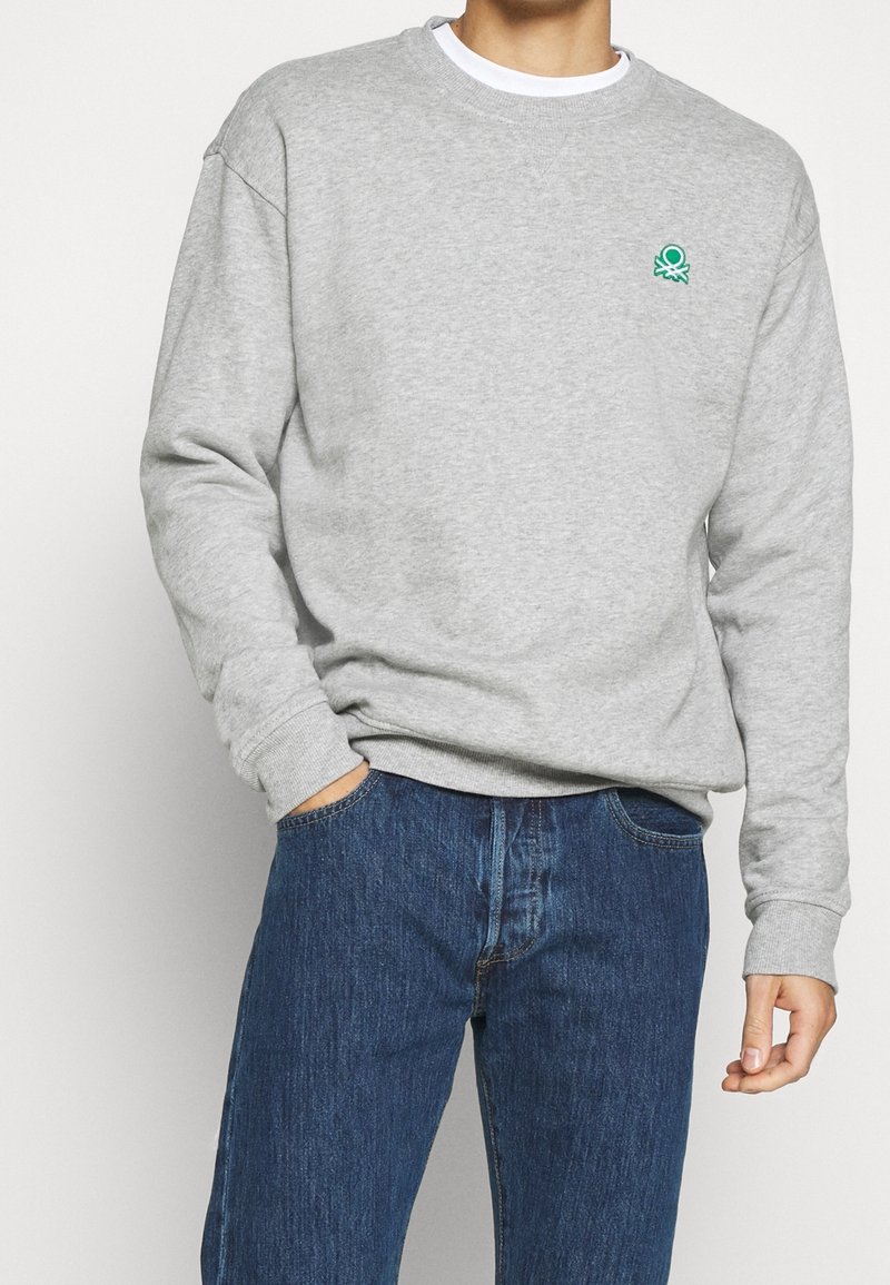 Grå sweatshirt i fleece med ribbade muddar och nederkant. Har en grön bläckfisklogga på vänster bröst. Kombineras med blå jeans.