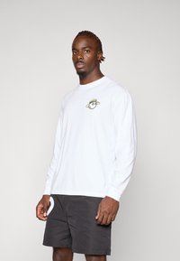 T-shirt bianca a maniche lunghe con un design colorato da cartone animato sul petto, abbinata a pantaloni corti scuri. Taglio semplice e casual.