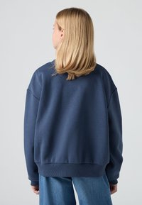 Felpa oversize blu navy con scollatura rotonda, polsini e bordo a costine, realizzata in tessuto di cotone testurizzato. Indossata con jeans chiari.