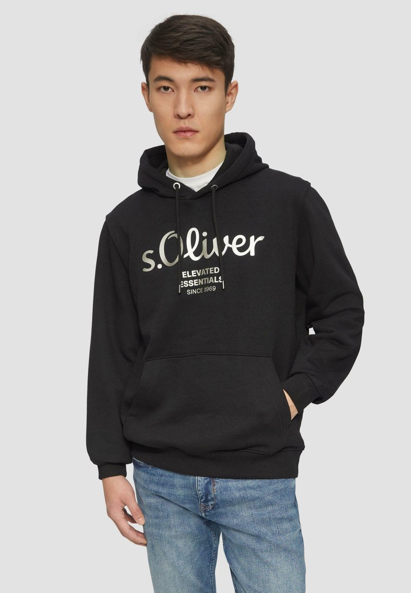 Zwarte hoodie met trekkoorden en een voorzak, met de tekst "s.Oliver Elevate Essentials since 1969" in het wit. Ggedragen met blauwe jeans.