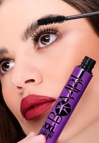 Confezione viola del mascara etichettata "BOMB! XXL INSTANT LENGTH" tenuta vicino al volto di un modello con lunghe ciglia scure e un colore di labbra rosso opaco.