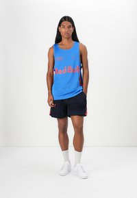 Blå mesh tanktop med "Red Bull"-logo, sorte shorts med røde accenter, hvite sneakers og grå sokker, båret av en modell mot en ensfarget bakgrunn.