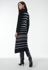 Lang zwarte jurk met horizontale witte strepen, lange mouwen, een aansluitend lijfje en een uitlopende rok. Gecombineerd met zwarte laarzen tot de knie.