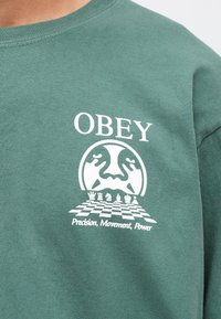 Zaļš kokvilnas T-krekls ar baltu grafiku, kas attēlo šaha dēli un tekstu "OBEY" kopā ar "Precizitāte, Kustība, Spēks."