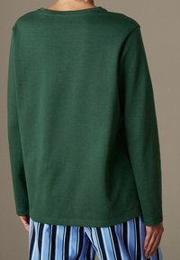 Eine Person trägt ein dunkelgrünes Langarmshirt und eine blau-weiß gestreifte Hose mit vertikalen Streifen. Sie steht mit dem Rücken zu einem neutralen Hintergrund.