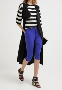 Un chaleco largo negro sobre un suéter a rayas en blanco y negro, combinado con unos pantalones cortos azules brillantes y unos tacones negros con tiras y tacón cuadrado.