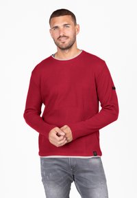 Chemise à manches longues rouge à côtes avec un col rond et un col intérieur gris contrastant. Présente une étiquette logo noire sur la manche gauche.