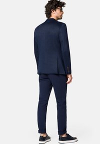 Granatowa marynarka o strukturalnym wzorze, w zestawie z dopasowanymi spodniami i teksturowanymi czarnymi trampkami. Do wyróżniających się detali należy brązowy akcent w postaci guzików.