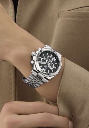 Sudraba nerūsējošā tērauda hronogrāfa pulkstenis ar melnu ciparnīcu, baltiem stundu rādītājiem un teksturētu rāmīti, ar tērauda saiti uz rokas siksnas.