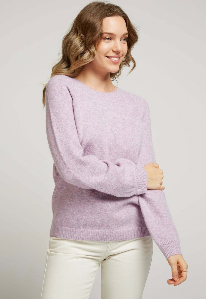 Pull léger violet en tissu doux et texturé. Il présente un col rond, des manches longues et un détail de bouton au niveau des poignets.