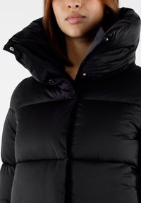 Giubbotto imbottito nero con colletto alto, caratterizzato da una chiusura con zip e un tessuto lucido. Nota per il suo design trapuntato e imbottito per il calore.