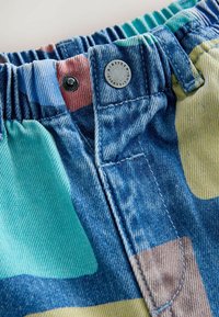 Primer plano de pantalones vaqueros multicolor con cintura elástica, botón metálico marcado con "NINETEEN EIGHTY" y detalles visibles de costura.