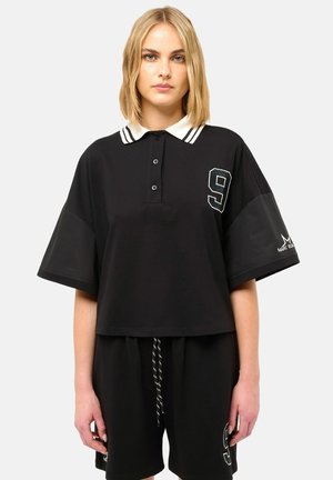 Femme blonde portant un polo noir ample avec un col à rayures blanches et un écusson numéro 9, associée à un short noir avec cordon de serrage.