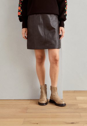 Frau trägt einen schwarzen Leder-Minirock, schwarzen Pullover mit bunten Blumenstickereien auf den Ärmeln und beigen Knöchelstiefeln, stehend auf einem Holzboden.