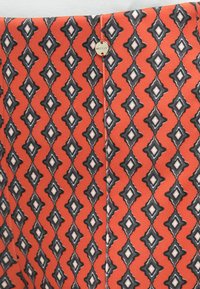 Rødt stof med diamantmønster i sort og hvid. Inkluderer et lille rundt metalmærke på siden. Glat tekstur.