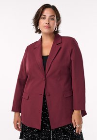 Kvinde med kort brunt hår iført sølvfarvede hoops, en mørkerød blazer over en sort top og en sort paljetteret nederdel mod en hvid baggrund.