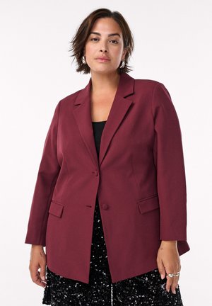 Žena s krátkymi hnedými vlasmi, ktorá má na sebe strieborné kruhové náušnice, tmavočervený blejzer cez čierny top a čiernu sukňu so flitrami na bielom pozadí.