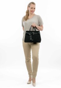 Sac à main noir au design structuré, avec une sangle texturée et un détail de boucle, porté par une personne vêtue d'un haut clair et d'un pantalon beige.