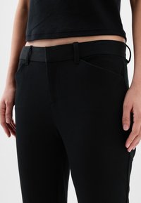 Taille et hanches d’une personne portant un pantalon noir ajusté avec passants de ceinture et un haut noir rentré, mains détendues le long du corps.