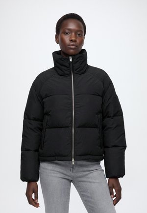 Person iført en sort dunjakke med høj krave, lynet op, og lysegrå jeans, stående mod en ensfarvet hvid baggrund.