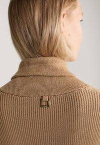 Beige gestrickter Pullover mit einem gerippten Muster, ausgestattet mit einem umgeschlagenen Kragen und einem kleinen goldenen Hardware-Detail auf der Rückseite.