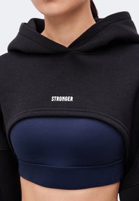Жена носи късо черно худи с текст "STRONGER" върху тъмносин спортен сутиен, показващ корема и шията.