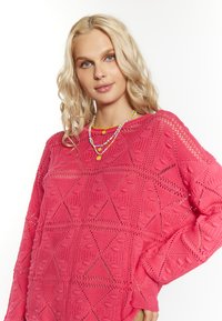 Rosa gestrickter Pullover mit geometrischem Muster, offenem Textur und langen Ärmeln. Geschichtete Perlenketten setzen farbige Akzente.