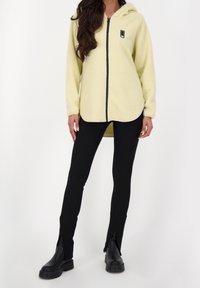 Gul fleecetröja med huva, dragkedja och svarta detaljer. Matchas med svarta leggings med slitsar vid nedre benet och kraftiga svarta boots.