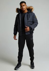 Jakna na napuhavanje tamno sive boje s kapuljačom obloženom krznom, više džepova i crnim detaljima, nošena preko crne majice i crnih trenirki.