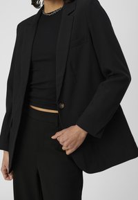 Blazer noir avec une fermeture à un seul bouton, des revers crantés et une coupe décontractée. Associé à un haut noir côtelé et un pantalon.