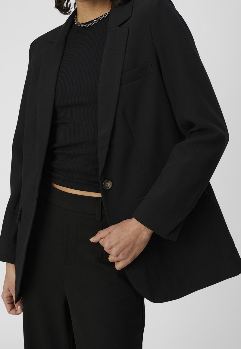 Blazer noir avec une fermeture à un seul bouton, des revers crantés et une coupe décontractée. Associé à un haut noir côtelé et un pantalon.