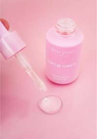 Fuktighetsgivende serum i en lys rosa flaske med pipette. Serumet er klart og samler seg i en dråpe på en rosa overflate. Teksten sier "IKKE VÆR TØRSTIG."