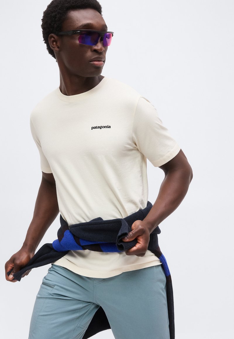 Hombre con camiseta beige Patagonia, pantalones azul grisáceo, gafas de sol oscuras, atando un suéter negro y azul alrededor de la cintura, mirando hacia su izquierda.