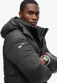 Chaqueta tipo puff en gris oscuro, con un patrón texturizado y una capucha ajustada. Incluye un parche de logo en la manga y detalles de cremallera visibles.