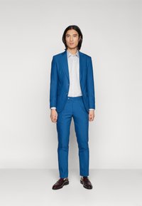 Modrý oblek na mieru s výstrihom do špicatého tvaru, slim fit nohavice a svetlo šedá košeľa. Nosí sa s lesklými tmavo hnedými topánkami.