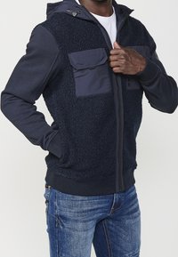 Marinblå huva-jacka med en mjuk, strukturerad kropp, två bröstfickor och ribbade muddar. Släta ärmar och dragkedjedetalj fulländar designen.