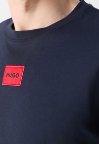 T-shirt en coton marine avec un col rond, présentant un patch rectangulaire brodé rouge avec le mot "HUGO" en noir.